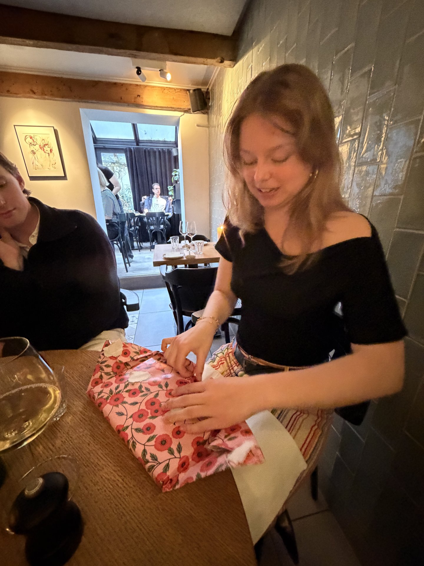 A woman is joyfully unwrapping a pink floral gift at a cozy restaurant on Govert Flinckstraat, Oude Pijp, Amsterdam